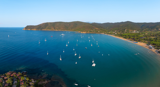 Aerial view of Lacona Beach with numerous sailboats and yachts anchored in the clear blue waters of the bay surrounded by lush green hills and coastal vegetation in Lacona Tuscany Italy.