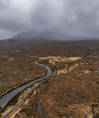 Aerial view of a winding river flowing through the rugged moorland landscape towards a lonely white cottage under misty mountains Isle of Skye Scotland United Kingdom. Aerial view of a winding river flowing through the rugged moorland landscape towards a lonely white cottage under misty mountains Isle of Skye Scotland United Kingdom.