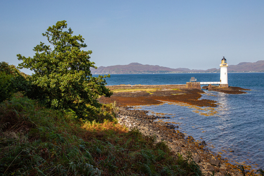 View of Tobermory Lighthouse with its white tower on a rocky shore lush green trees in the foreground and distant hills across the blue sea Isle of Mull Scotland United Kingdom. View of Tobermory Lighthouse with its white tower on a rocky shore lush green trees in the foreground and distant hills across the blue sea Isle of Mull Scotland United Kingdom.