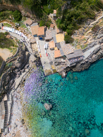 Aerial view of Cala Deia with stone buildings small boats on a ramp and people by the turquoise water on the coast of Spain. Aerial view of Cala Deia with stone buildings small boats on a ramp and people by the turquoise water on the coast of Spain.