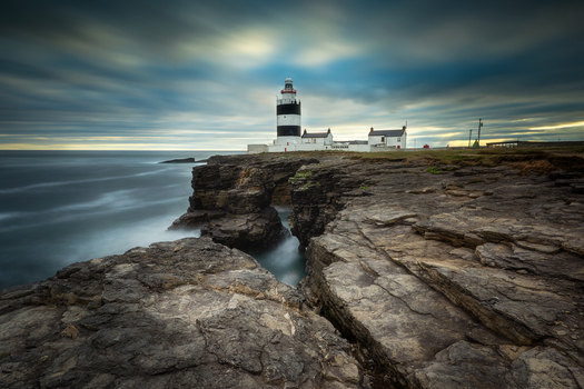 View of Hook Lighthouse standing on rugged rocky cliffs under a dramatic cloudy sky with long exposure sea waves County Wexford Ireland. View of Hook Lighthouse standing on rugged rocky cliffs under a dramatic cloudy sky with long exposure sea waves County Wexford Ireland.