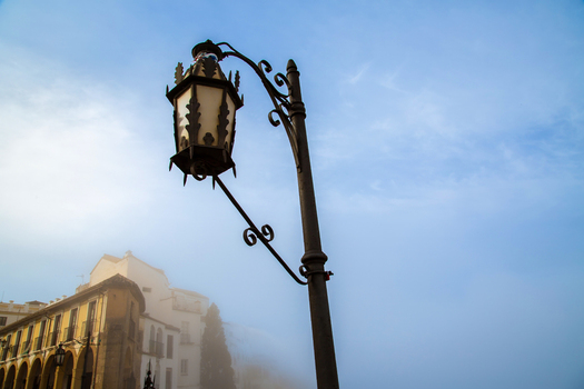 View of a decorative wrought iron street lamp with historic white buildings and arched arcades partially obscured by morning mist in Ronda Andalusia Spain.