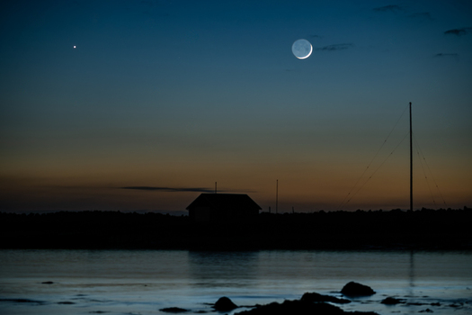 View of the crescent moon and a bright star shining in the twilight sky over a silhouetted building and calm water at Seltjarnarnes Seltjarnarnesbaer Iceland. View of the crescent moon and a bright star shining in the twilight sky over a silhouetted building and calm water at Seltjarnarnes Seltjarnarnesbaer Iceland.