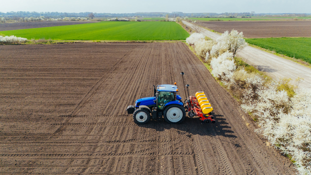 Aerial view of a blue tractor working the dark soil of a field juxtaposed against vibrant green fields and white blossoming trees Sremska Mitrovica Vojvodina Serbia. Aerial view of a blue tractor working the dark soil of a field juxtaposed against vibrant green fields and white blossoming trees Sremska Mitrovica Vojvodina Serbia.