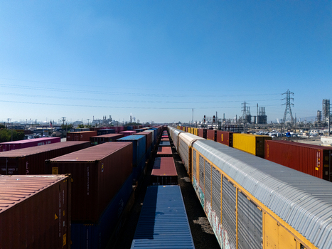Aerial view of a freight train with colorful shipping containers and railcars on tracks in an industrial area under a clear blue sky in Carson California United States.
