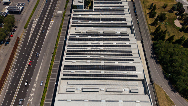 Aerial view of a large industrial warehouse roof covered with solar panels situated next to a busy highway with moving vehicles in Milan Lombardy Italy. Aerial view of a large industrial warehouse roof covered with solar panels situated next to a busy highway with moving vehicles in Milan Lombardy Italy.