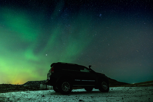 View of a dark SUV parked on the snowy ground under the mesmerizing green aurora borealis and starry sky Krysuvikurkirkja Hafnarfjardarkaupstadur Iceland.
