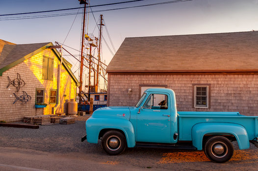 View of a vintage blue pickup truck with a dog in the driver s seat parked by shingled coastal buildings and fishing boat masts in Edgartown Massachusetts United States.