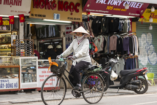 Hanoi Vietnam 11 November 2025 View of a woman wearing a traditional conical hat riding a bicycle past busy shops in the historic Old Quarter.