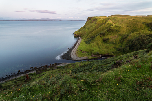 View of the rugged green cliffs and calm sea of the Isle of Skye coastline with stone ruins on the grassy slopes Highland Council Scotland United Kingdom.