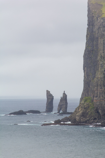 View of the iconic Risin og Kellingin sea stacks rising from the North Atlantic Ocean near steep coastal cliffs under a cloudy sky in the Faroe Islands.