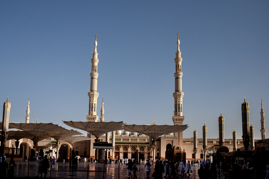 View of Al Masjid e Nabawi with its iconic minarets and large retractable umbrellas in the courtyard with people walking in the foreground in Madinah Al Madinah Province Saudi Arabia. View of Al Masjid e Nabawi with its iconic minarets and large retractable umbrellas in the courtyard with people walking in the foreground in Madinah Al Madinah Province Saudi Arabia.