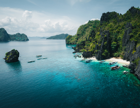 Aerial view of dramatic limestone cliffs turquoise waters and traditional outrigger boats anchored near a white sand beach in El Nido Mimaropa Philippines. Aerial view of dramatic limestone cliffs turquoise waters and traditional outrigger boats anchored near a white sand beach in El Nido Mimaropa Philippines.
