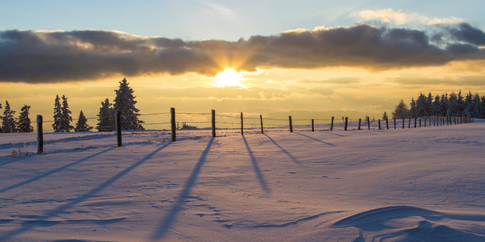 View of the sun bursts through clouds casting long shadows from a fence across a snow covered field lined with trees Schöckl Austria. View of the sun bursts through clouds casting long shadows from a fence across a snow covered field lined with trees Schöckl Austria.