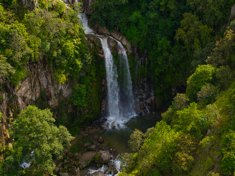 Aerial view of a cascading waterfall flowing into a pool within a lush forested canyon in Nepal.