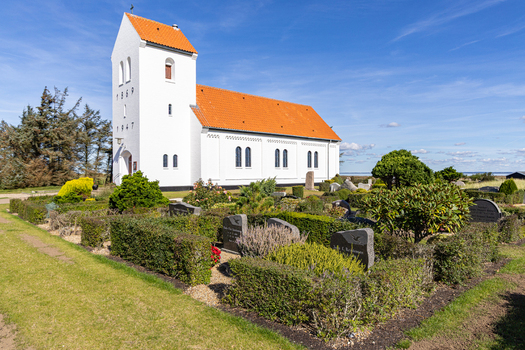 View of Haurvig Kirke with its white facade and orange tiled roof surrounded by a well kept churchyard under a clear blue sky in Hvide Sande Denmark. View of Haurvig Kirke with its white facade and orange tiled roof surrounded by a well kept churchyard under a clear blue sky in Hvide Sande Denmark.