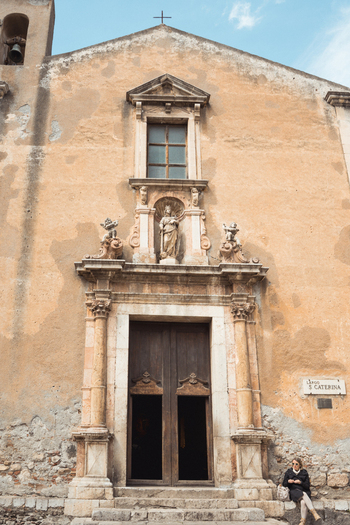 View of the weathered facade of a historic church with a bell tower a statue above the door and a cross on top Taormina Catania Italy.