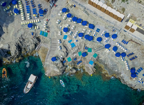 Aerial view of a rocky beach club with blue umbrellas sunbeds and boats in turquoise water Capri Campania Italy.