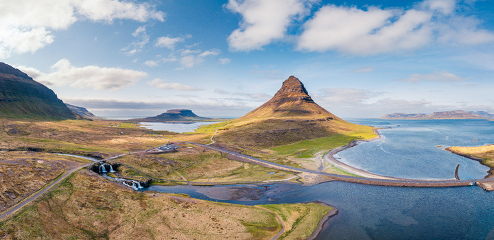 Aerial view of Kirkjufell mountain surrounded by green fields a winding road and the coastline under a blue sky in Iceland. Aerial view of Kirkjufell mountain surrounded by green fields a winding road and the coastline under a blue sky in Iceland.
