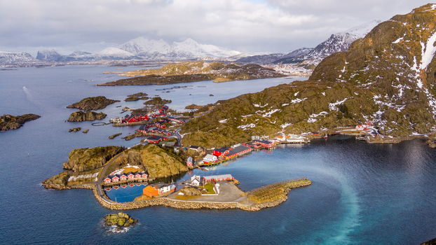 Aerial view of the Lofoten archipelago featuring traditional red wooden houses rugged rocky coastline and snow capped mountains in Nordland Norway.