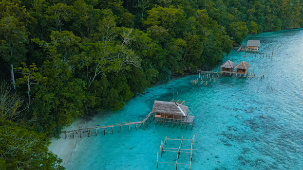 Aerial view of overwater bungalows nestled along a lush tropical coastline with vibrant turquoise waters in Raja Ampat Southwest Papua Indonesia.
