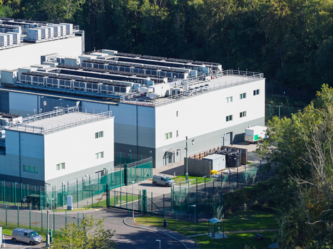 Aerial view of a modern data center facility with rooftop HVAC units perimeter fencing and surrounding trees in the United Kingdom.
