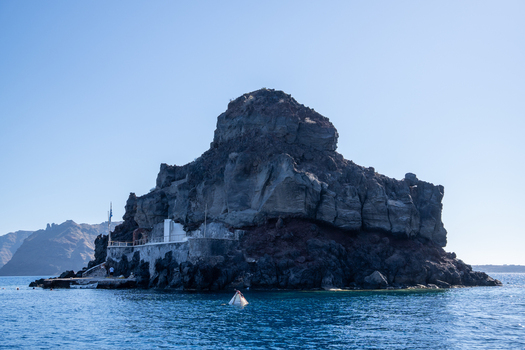 View of the Skaros Rock volcanic formation with a small white structure at its base under a clear blue sky in Fira Greece. View of the Skaros Rock volcanic formation with a small white structure at its base under a clear blue sky in Fira Greece.