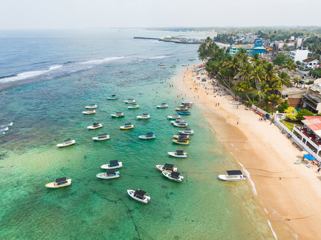 Aerial view of the turquoise coastline with numerous boats anchored near the sandy beach and palm trees in Hikkaduwa Southern Province Sri Lanka. Aerial view of the turquoise coastline with numerous boats anchored near the sandy beach and palm trees in Hikkaduwa Southern Province Sri Lanka.