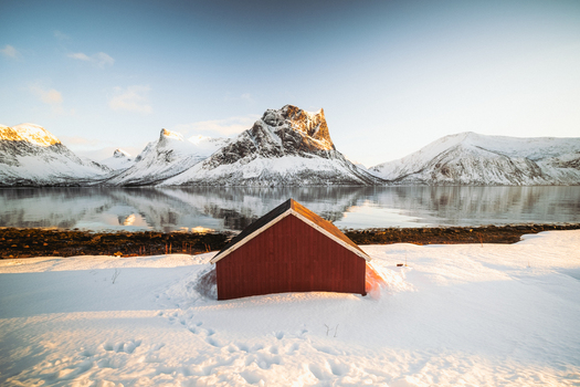 View of a solitary red cabin stands bravely against the snowy foreground facing a majestic mountain range reflected in the calm waters Indregård Troms Norway. View of a solitary red cabin stands bravely against the snowy foreground facing a majestic mountain range reflected in the calm waters Indregård Troms Norway.