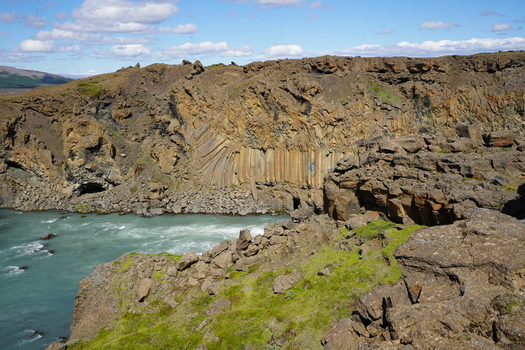 View of the dramatic basalt column rock formations along the river canyon with mossy green slopes under a blue sky in Thingeyri Isafjardarbaer Iceland. View of the dramatic basalt column rock formations along the river canyon with mossy green slopes under a blue sky in Thingeyri Isafjardarbaer Iceland.