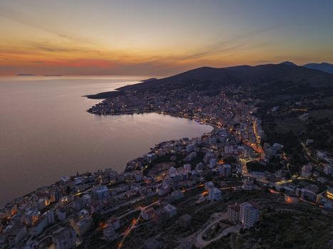 Aerial view of Saranda at sunset featuring the coastal city lights buildings along the bay and surrounding hills in Saranda Vlore County Albania. Aerial view of Saranda at sunset featuring the coastal city lights buildings along the bay and surrounding hills in Saranda Vlore County Albania.