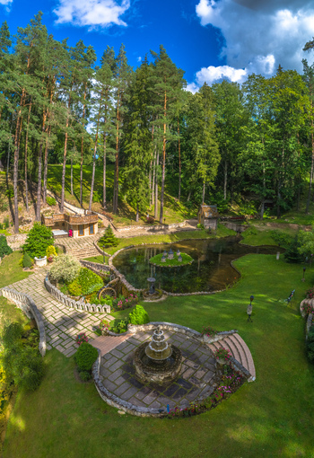 Aerial view of a lush park featuring a tranquil pond with a small island a stone fountain and manicured gardens surrounded by tall pine trees in Latvia. Aerial view of a lush park featuring a tranquil pond with a small island a stone fountain and manicured gardens surrounded by tall pine trees in Latvia.