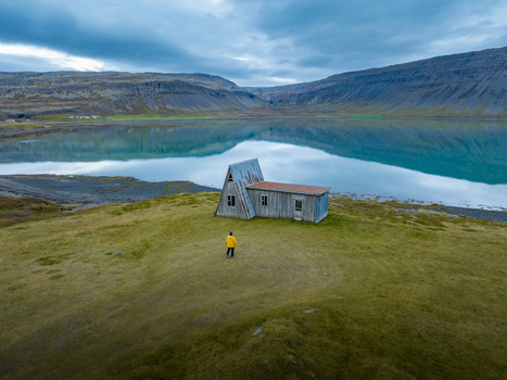 Aerial view of a lone figure standing by a weathered building on a grassy peninsula with mirror like waters reflecting the brooding sky and majestic mountains Westfjords Iceland. Aerial view of a lone figure standing by a weathered building on a grassy peninsula with mirror like waters reflecting the brooding sky and majestic mountains Westfjords Iceland.