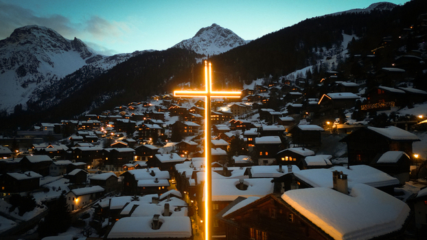 Aerial view of the illuminated cross glows warmly against the snow dusted rooftops and dark silhouettes of the towering Alps Grimentz Valais Switzerland.