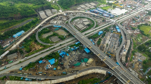 Aerial view of the JNPT Interchange featuring complex highway ramps shipping container yards and industrial infrastructure surrounded by lush green hills in Navi Mumbai Maharashtra India. Aerial view of the JNPT Interchange featuring complex highway ramps shipping container yards and industrial infrastructure surrounded by lush green hills in Navi Mumbai Maharashtra India.
