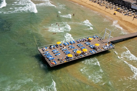 Aerial view of a wooden pier extending into the sea featuring rows of blue sun loungers and yellow umbrellas with people swimming near the beach in Kumkoy Istanbul Turkey. Aerial view of a wooden pier extending into the sea featuring rows of blue sun loungers and yellow umbrellas with people swimming near the beach in Kumkoy Istanbul Turkey.
