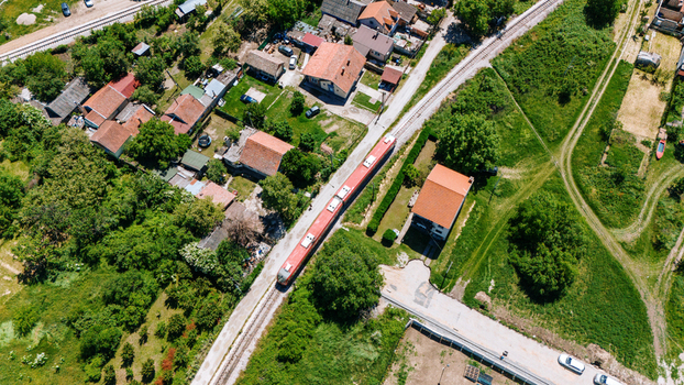 Aerial view of a red passenger train traveling on railway tracks through a suburban neighborhood with red roofed houses and lush green gardens Pancevo Vojvodina Serbia.