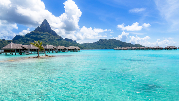 View of overwater bungalows in the turquoise lagoon with Mount Otemanu in the background under a blue sky with white clouds Bora Bora Leeward Islands French Polynesia.