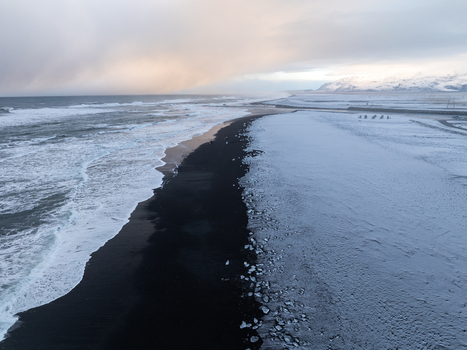 Aerial view of a dramatic black sand beach featuring snow covered shores crashing ocean waves and a vast cloudy sky over the rugged coastline of Iceland. Aerial view of a dramatic black sand beach featuring snow covered shores crashing ocean waves and a vast cloudy sky over the rugged coastline of Iceland.