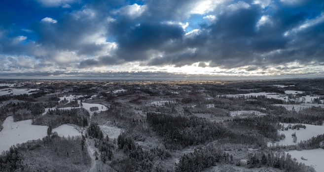 Aerial view of a snow covered landscape featuring dense forests and open fields under a dramatic cloudy sky with sunlight breaking through Latvia. Aerial view of a snow covered landscape featuring dense forests and open fields under a dramatic cloudy sky with sunlight breaking through Latvia.