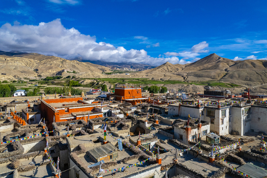 Aerial view of a traditional Tibetan monastery with orange and white buildings adorned with prayer flags set against a backdrop of arid mountains in Nepal.