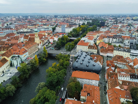 Aerial view of the Kunsthaus Graz and the Mur River flowing through the historic city center with red tiled rooftops and lush trees in Graz Styria Austria.