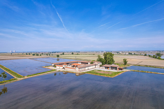 Aerial view of a traditional farm complex surrounded by flooded rice fields under a clear blue sky in Trino Piedmont Italy.