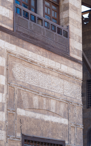 View of the intricate Arabic calligraphy and stone carvings on the facade of the Al Ghuri Dome complex with a traditional wooden balcony in Cairo Egypt. View of the intricate Arabic calligraphy and stone carvings on the facade of the Al Ghuri Dome complex with a traditional wooden balcony in Cairo Egypt.