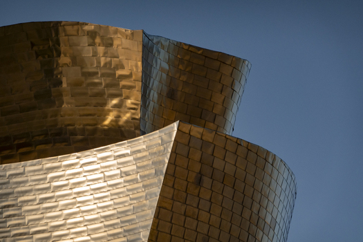 View of the curved titanium panels of the Guggenheim Museum Bilbao reflecting golden light against a clear blue sky Bilbao Euskadi Spain. View of the curved titanium panels of the Guggenheim Museum Bilbao reflecting golden light against a clear blue sky Bilbao Euskadi Spain.