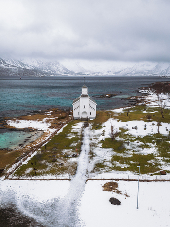 Aerial view of the pristine white Gimsøy Church stands in stark contrast against the muted blues of the surrounding waters Gimsøysand Nordland Norway. Aerial view of the pristine white Gimsøy Church stands in stark contrast against the muted blues of the surrounding waters Gimsøysand Nordland Norway.