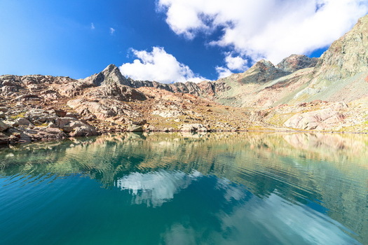 View of a crystal clear alpine lake reflecting rugged mountain peaks and white clouds under a bright blue sky in the Italian Alps near Chiesa in Valmalenco Lombardy Italy. View of a crystal clear alpine lake reflecting rugged mountain peaks and white clouds under a bright blue sky in the Italian Alps near Chiesa in Valmalenco Lombardy Italy.