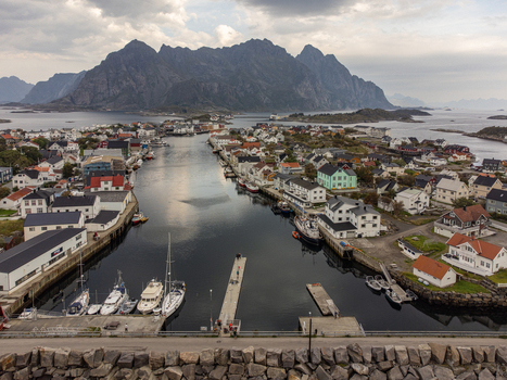 Aerial view of calm waters reflecting the quaint village architecture nestled beneath the majestic mountain ranges Henningsvær Nordland Norway. Aerial view of calm waters reflecting the quaint village architecture nestled beneath the majestic mountain ranges Henningsvær Nordland Norway.
