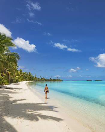 View of a woman walking along the white sand beach of One Foot Island in Aitutaki with turquoise lagoon waters lush palm trees and a clear blue sky in Arutanga Cook Islands. View of a woman walking along the white sand beach of One Foot Island in Aitutaki with turquoise lagoon waters lush palm trees and a clear blue sky in Arutanga Cook Islands.