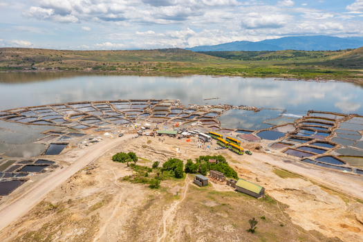 Aerial view of Lake Katwe salt pans with workers and vehicles surrounded by arid landscape and water under a cloudy sky in Kasese Western Region Uganda.